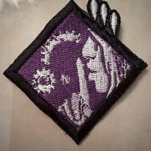 Puede incluir: Un parche bordado en forma de diamante con fondo morado, un diseño gráfico blanco de una figura estilizada y tres marcas de garras que sobresalen de la esquina superior derecha, todo bordeado por un grueso borde negro cosido.