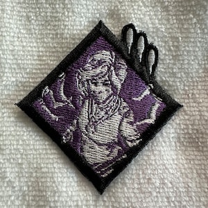 Puede incluir: Parche bordado con una figura estilizada en blanco y morado, enmarcada en un diamante negro. El diseño incluye un rostro detallado y brazos extendidos, con un elemento único de tres lóbulos en la parte superior.