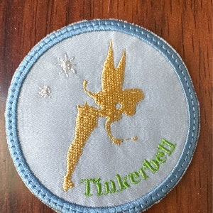 May include: Embroidered patch featuring Tinkerbell in gold against a light blue background. The patch has a light blue border with green text that reads "Tinkerbell". Two small white stars are also embroidered on the patch.