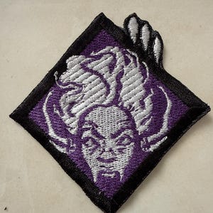 May include: An embroidered patch in a diamond shape, featuring a stylized face with flowing hair. The design uses purple, white, and black thread. The patch has a black border and is likely for clothing or accessories.