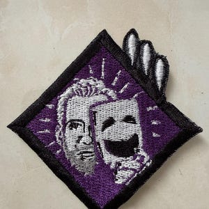 May include: Embroidered patch featuring a purple diamond shape with a black border. The patch depicts two theatrical masks, one smiling and one frowning, in white thread against a purple background. A hand of white thread is visible.