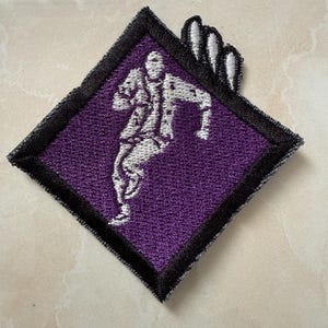 May include: Embroidered patch featuring a figure in motion, rendered in white thread against a deep purple background. The patch has a black border and a unique shape, with a stylized hand design.