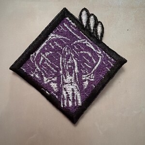 以下が含まれることがあります： 紫色の背景に、腕を上げた人物の白い刺繍グラフィックが施された、ひし形の刺繍ワッペン。ワッペンには太い黒の縁取りがあり、右上隅から3つの小さな白い爪のような形が突き出ている。