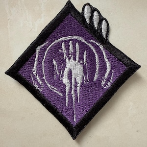 May include: Embroidered patch with a purple diamond shape and a black border. The design features a white skeletal hand reaching out from a circle. The patch is likely for clothing or accessories.