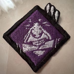 May include: A diamond-shaped embroidered patch featuring a purple background with a white graphic of a figure holding their head in their hands. The patch has a thick black border and a small white wing-like detail extending from the top right corner.