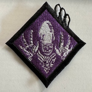 Könnte beinhalten: Ein lila-schwarzer, gestickter Aufnäher in Diamantenform. Der Aufnäher zeigt ein weißes Skelett-Design mit ausgestreckten Armen und einem Schädel. Der Aufnäher hat einen schwarzen Rand und ein weißes Handdesign.