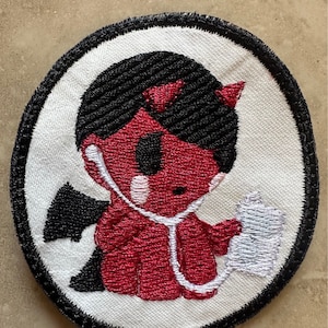 May include: Embroidered patch featuring a red devil character with black hair, horns, and wings, wearing a stethoscope. The devil is holding a medical chart. The patch has a white background with a black border.