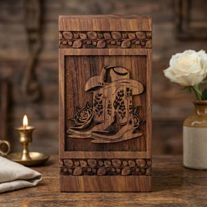 Op de afbeelding: Een rechthoekige houten kist met een gesneden ontwerp met een cowboyhoed, laarzen en rozen. De kist heeft een donkerbruine afwerking en is versierd met een bloemenrand. Het ontwerp heeft een rustiek, country-western thema.