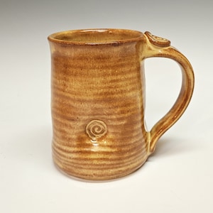 May include: A handmade ceramic mug with a brown glaze and a swirl design. The mug has a rounded body and a handle.