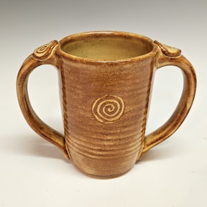 May include: A brown ceramic mug with two handles and a spiral design. The mug has a light brown glaze.