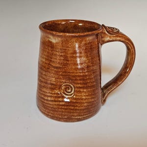 May include: A brown ceramic mug with a textured surface and a white swirl design. The mug has a handle with a white swirl design.
