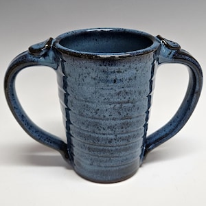 May include: A ceramic mug featuring a deep blue speckled glaze with a ribbed, cylindrical body and two symmetrical handles on opposite sides. The rim and handle accents are finished in a darker, glossy tone, creating a textured appearance.