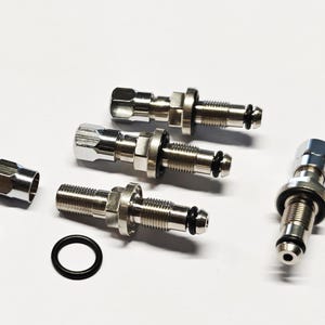 Op de afbeelding: Vier zilveren metalen fittingen met zwarte rubberen O-ringen. De fittingen hebben een schroefdraad einde en een recht einde. Eén fitting heeft een zeskantige moer aan het schroefdraad einde.