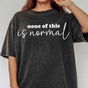 Puede incluir: Camiseta gris oscuro con la frase blanca "none of this is normal". La camiseta tiene un aspecto vintage y desgastado. El texto está centrado en la parte delantera de la prenda, con una fuente sencilla y elegante.