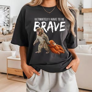 Op de afbeelding: Zwart T-shirt met de witte tekst "ULTIMATELY I HAVE TO BE BRAVE". Het shirt heeft een afbeelding van twee apen, een grijze en een oranje. Het model draagt een lichtgrijze broek.