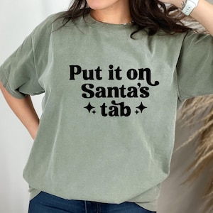 Puede incluir: Camiseta verde salvia con la frase "Put it on Santa's tab" en negro, con letras retro y estrellas. La camiseta tiene un corte holgado y un estilo casual.