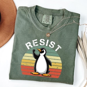 Puede incluir: Una camiseta verde con un gráfico de estilo vintage de un pingüino levantando su puño en el aire. El gráfico está rodeado por una puesta de sol roja, naranja y amarilla. El texto "RESIST" está escrito en blanco sobre el gráfico.