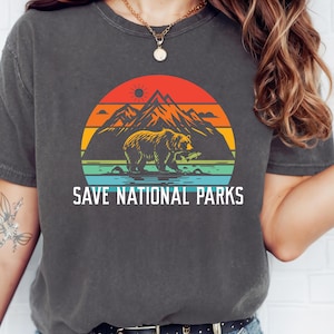 Puede incluir: Una camiseta gris oscuro con un gráfico de estilo vintage de un oso pescando en un lago con montañas en el fondo. El gráfico está rodeado por un círculo de colores arcoíris. El texto "SAVE NATIONAL PARKS" está impreso debajo del gráfico.