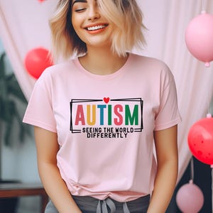 Puede incluir: Una camiseta rosa con el texto "AUTISMO - VER EL MUNDO DE FORMA DIFERENTE" en letras de colores con un pequeño corazón rojo sobre la palabra "AUTISMO".