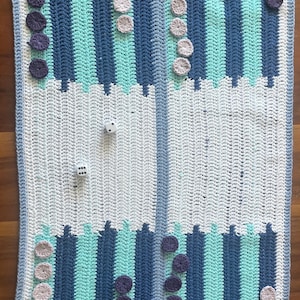Pode incluir: Um tabuleiro de gamão de crochê feito à mão em tons de azul, verde-azulado e branco. O tabuleiro apresenta seções listradas e uma borda, com peças roxas e rosa claro e dois dados.