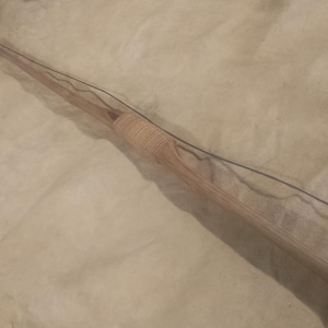 Pode incluir: Um arco longo de madeira com uma corda escura. O arco é castanho claro com uma pega enrolada. O arco está sobre uma superfície de cor clara.