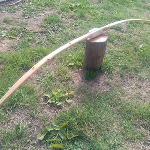 May include: A longbow crafted from light-coloured wood with a smooth, curved design. The centre grip area is wrapped in a light-coloured material, providing a distinct contrast to the natural grain of the wooden limbs that taper towards each end.