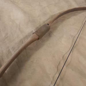 Pode incluir: Um arco de madeira com uma corda escura, repousando sobre uma superfície clara. O arco tem um design curvo com uma pega no centro. O arco é feito de madeira de cor clara.