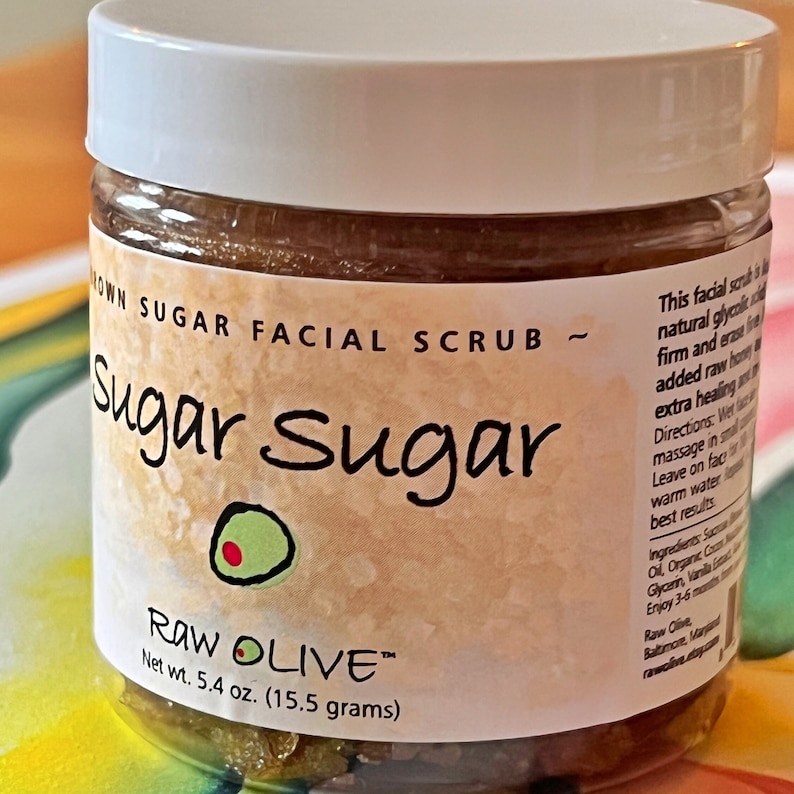 Close up of a clear jar with a white lid that reads Sugar Sugar, Brown Sugar Facial Scrub, Raw Olive, moisturing and exfoliating natural scrub