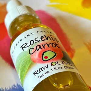 Close-up of a glass bottle of Raw Olive Rosehip Carrot antioxidant facial serum. The label is white with a redish pink rosehip, and a green olive.