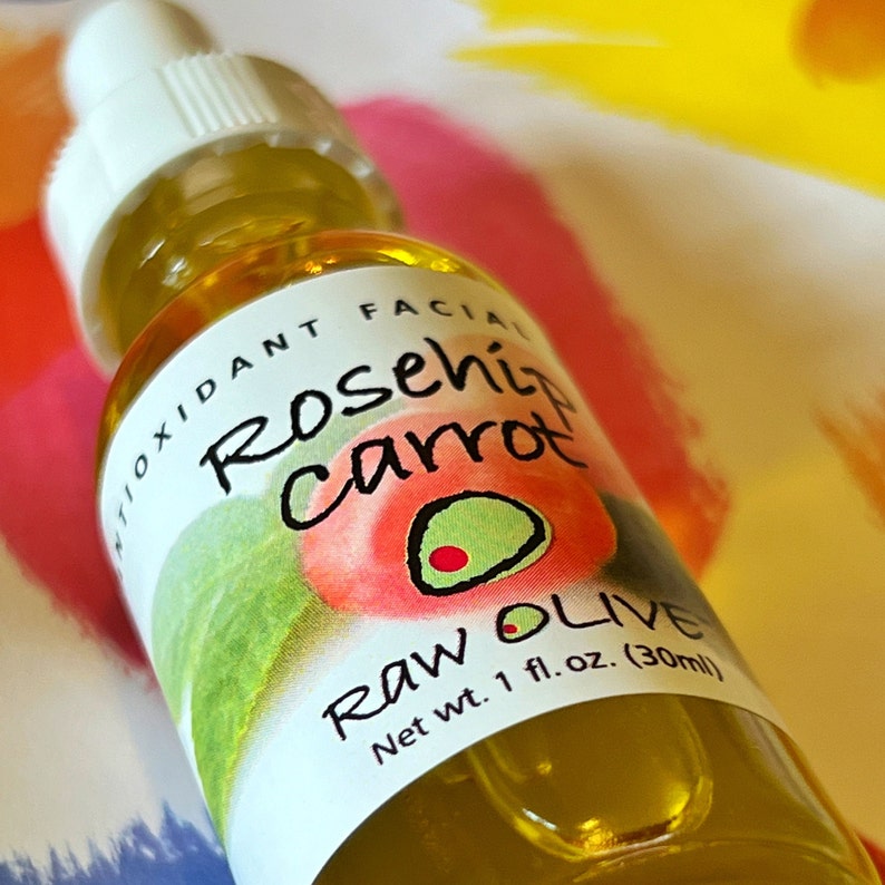Close-up of a glass bottle of Raw Olive Rosehip Carrot antioxidant facial serum. The label is white with a redish pink rosehip, and a green olive.