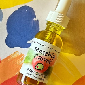 Close-up of a glass bottle of Raw Olive Rosehip Carrot antioxidant facial serum on a colorful background. The label is white with a redish pink rosehip, and a green olive.