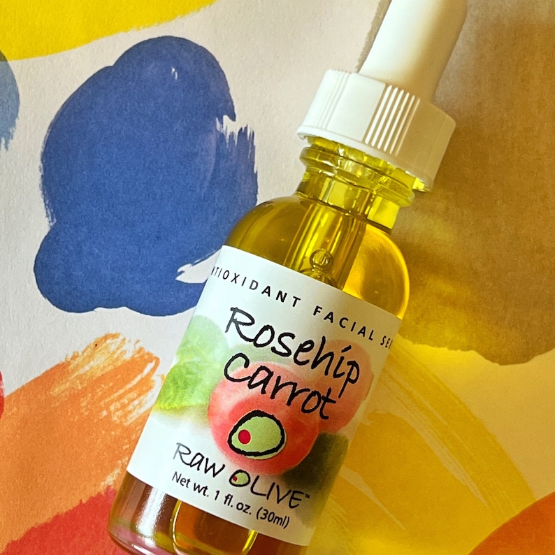 Close-up of a glass bottle of Raw Olive Rosehip Carrot antioxidant facial serum on a colorful background. The label is white with a redish pink rosehip, and a green olive.