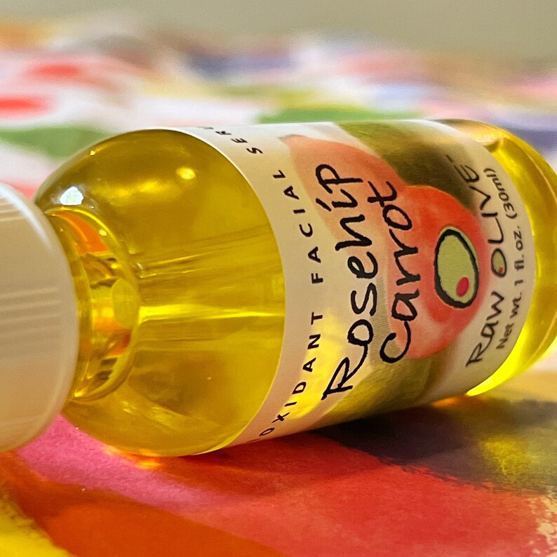Close-up of a glass bottle of Raw Olive Rosehip Carrot antioxidant facial serum lying on its side. The label is white with a redish pink rosehip, and a green olive.