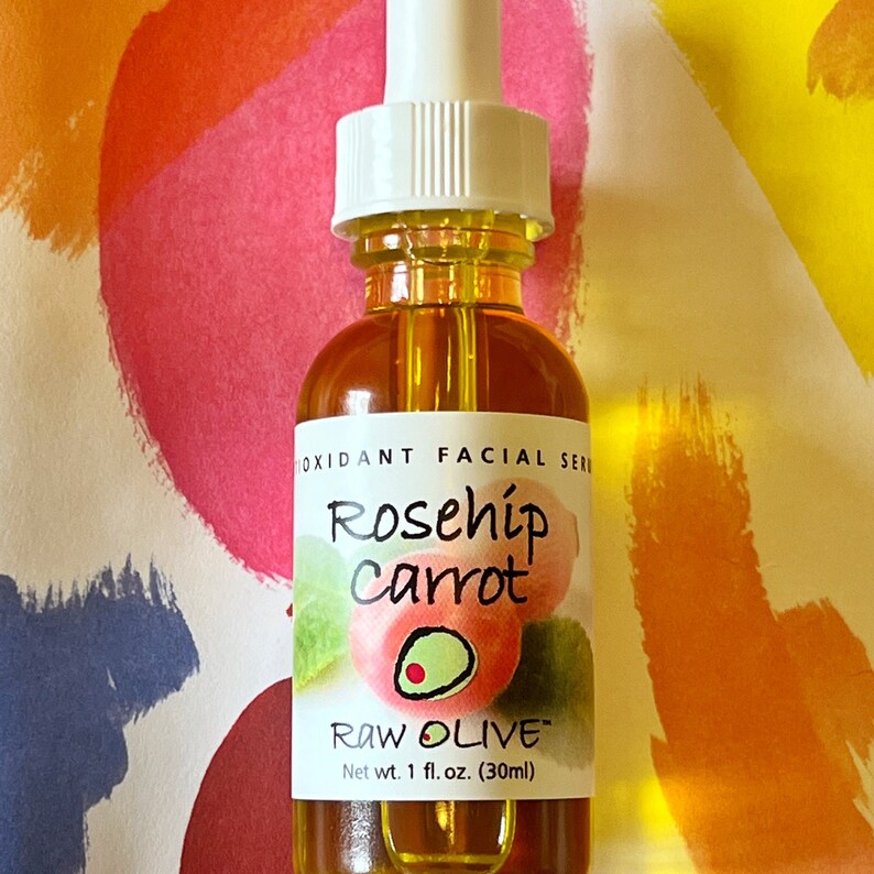 Close-up of a glass bottle of Raw Olive Rosehip Carrot antioxidant facial serum. The label is white with a redish pink rosehip, and a green olive.