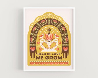 May include: A framed art print featuring a stylized floral design in shades of yellow, brown, and red. The artwork includes the text "HELD IN LOVE WE GROW" and decorative elements like birds and hearts. The print is set against a light background.