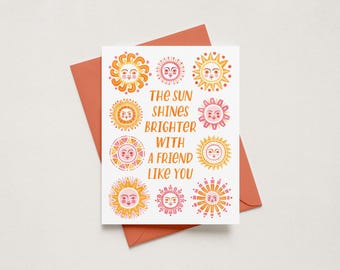 May include: A white greeting card with a coral envelope featuring the text THE SUN SHINES BRIGHTER WITH A FRIEND LIKE YOU in orange letters, surrounded by nine distinct illustrations of suns with faces in shades of yellow, orange, and pink.
