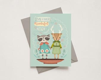 May include: A greeting card with a light blue background featuring a raccoon and a deer standing in a bowl. The raccoon wears a blue dress with white flowers, and the deer wears a green dress. The text reads, You're a wildly wonderful friend.