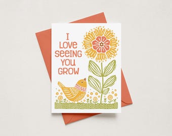 May include: A white greeting card with a coral envelope featuring a yellow bird and a large orange and yellow flower. The text reads I LOVE SEEING YOU GROW in orange letters. The design includes small green leaves and flowers on a dotted ground.