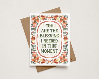 May include: A white greeting card with a brown envelope featuring the text YOU ARE THE BLESSING I NEEDED IN THIS MOMENT in dark green letters. The card is decorated with a symmetrical border of orange birds, red flowers, and green leaves.