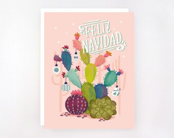 May include: A greeting card with a pink background features a festive cactus illustration. The cactus is decorated with ornaments and topped with the words "Feliz Navidad" in white script. The card has a white border.
