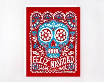 May include: A red greeting card with a detailed illustration of a blue and white sugar skull. The skull design is surrounded by floral and decorative elements. The words "Feliz Navidad" are displayed in white script.