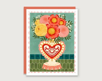 May include: A bouquet of pink, orange, and yellow flowers in a white heart-shaped vase with red and white details. The vase is sitting on a green and white tiled surface.