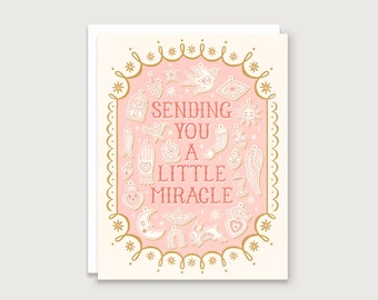 May include: A pink greeting card with a gold frame and white illustrations of charms and symbols. The text inside the frame reads "Sending you a little miracle".