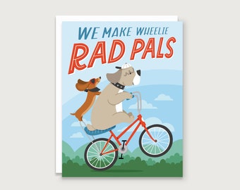 May include: A greeting card with a cartoon illustration of two dogs riding a bicycle. The text on the card reads "We make wheelie rad pals".