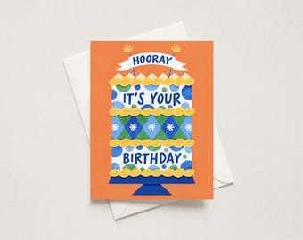 May include: An orange greeting card featuring a multi-tiered cake illustration with blue and green polka dots and argyle patterns. The text reads HOORAY IT'S YOUR BIRTHDAY in blue letters. The cake sits on a dark blue stand with yellow frosting accents.