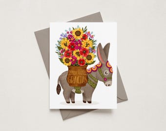 May include: A greeting card featuring a gray donkey carrying a brown basket filled with a vibrant bouquet of sunflowers, red poppies, and blue wildflowers. The basket has the word GRACIAS printed on the front, and the donkey wears a colorful harness.