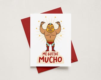 May include: A white greeting card featuring a cartoon illustration of a muscular luchador wrestler wearing a yellow mask, red trunks, and red boots. The wrestler is surrounded by yellow stars and the text ME GUSTAS MUCHO in red letters.