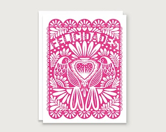 May include: A rectangular greeting card featuring a vibrant pink and white paper-cut style design. The illustration displays two birds facing a central heart, surrounded by floral patterns and the word FELICIDADES printed in a curved arch.