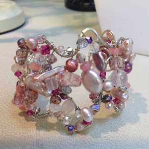 May include: A silver wire bracelet with pink, white, and purple beads. The bracelet is made with a variety of beads, including pearls, crystals, and gemstones.