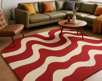 May include: A red and cream-colored area rug with a wavy pattern dominates the foreground. The rug is placed in a living room setting, with a green leather sectional sofa, a wooden coffee table, and a mid-century modern chair. The room is decorated with throw pillows and a floor lamp.